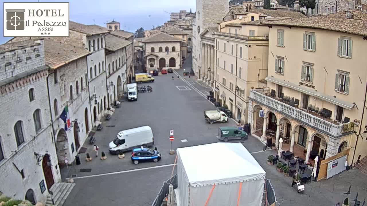 A mostly empty town square in Assisi, Italy, is seen on an overcast day with several vehicles parked and a few people walking around.