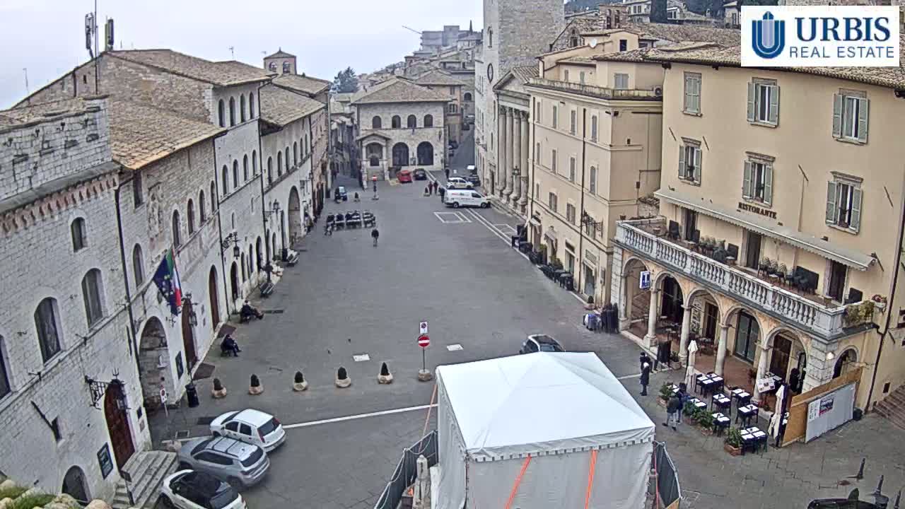 A mostly empty town square in overcast weather is surrounded by stone buildings, with a few parked cars and pedestrians visible.