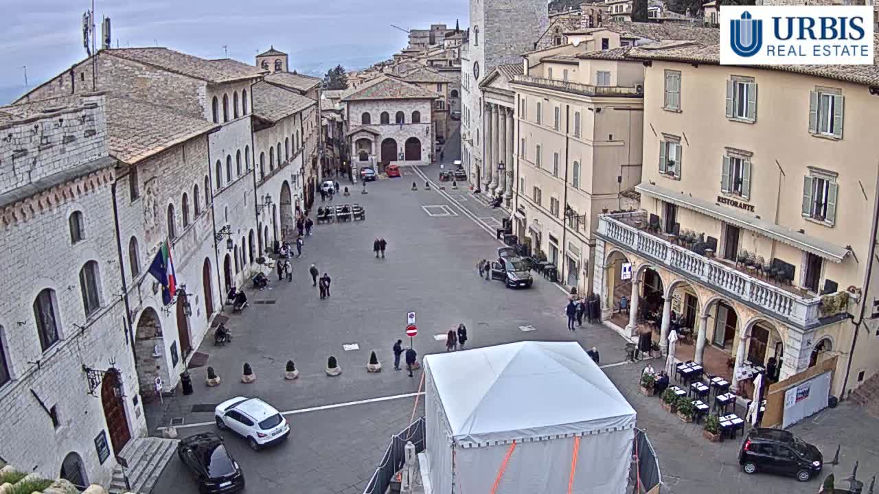A mostly empty town square in a historic Italian town on an overcast day, with several parked cars and a few pedestrians.