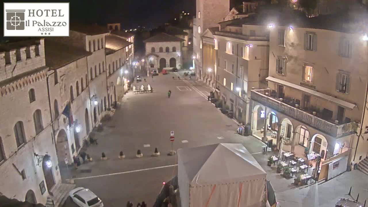 A mostly empty town square at night, surrounded by old buildings, is illuminated by streetlights.