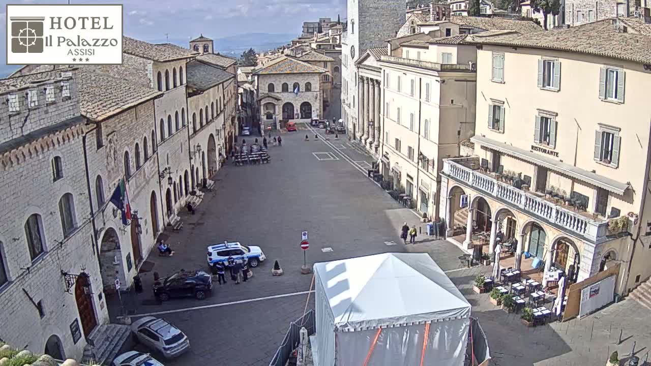 A large, mostly empty town square in bright daylight is surrounded by stone buildings, with a few cars, people, and a large white tent visible.