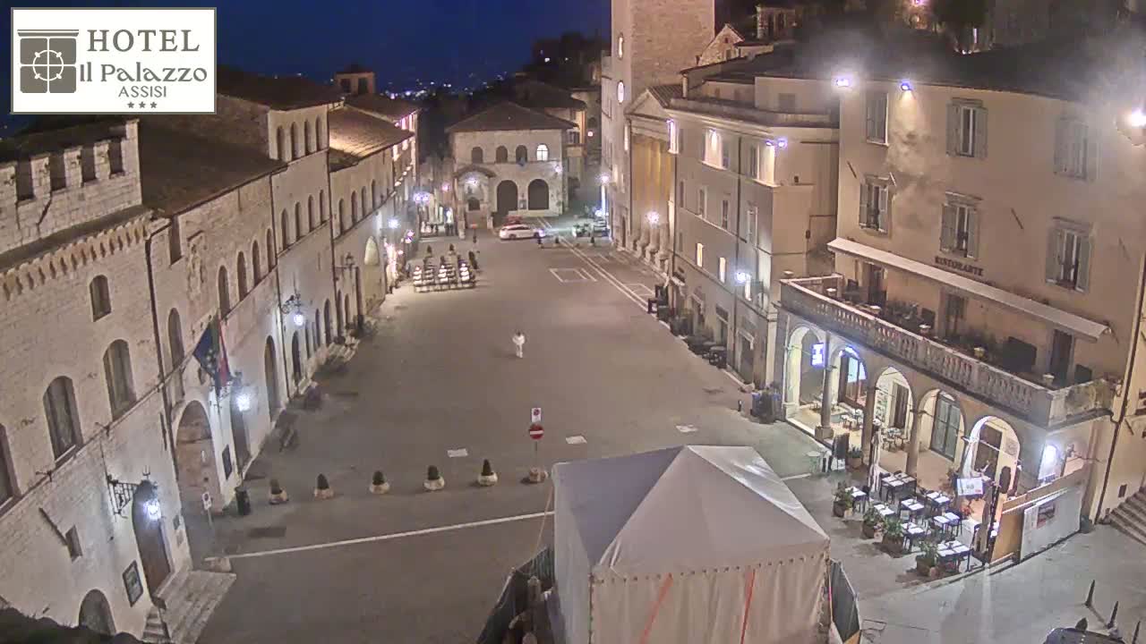 An empty, stone plaza at night, surrounded by illuminated buildings, with a small white tent in the foreground.