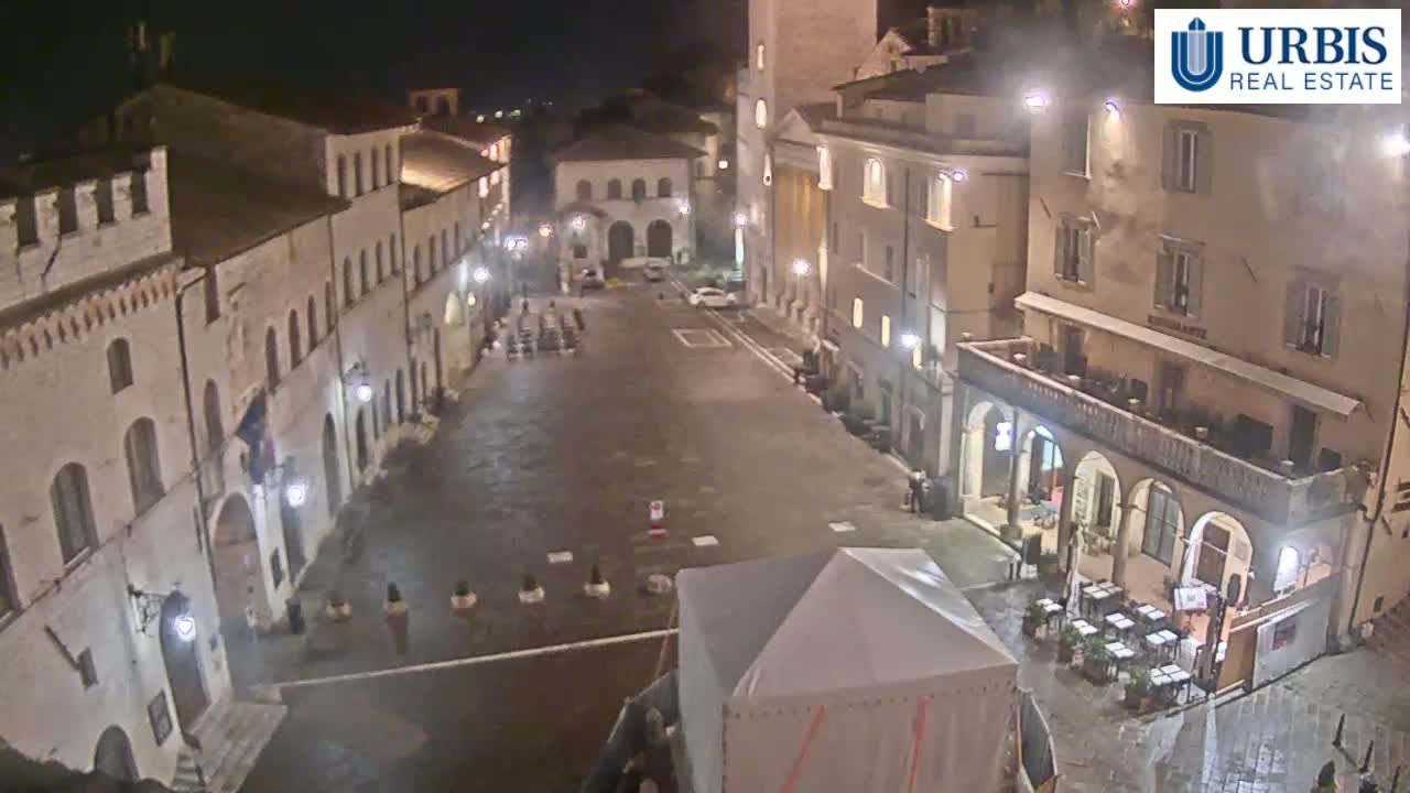 A nighttime view of a mostly empty town square surrounded by buildings, with a small tent in the foreground and a few outdoor tables and chairs visible near one building.