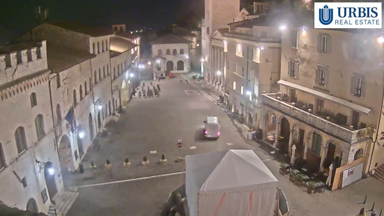 A mostly empty town square at night, surrounded by historic buildings, with a single van driving through.