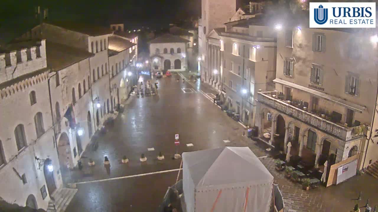 A wet, empty town square at night is surrounded by several multi-story buildings lit by street lamps.