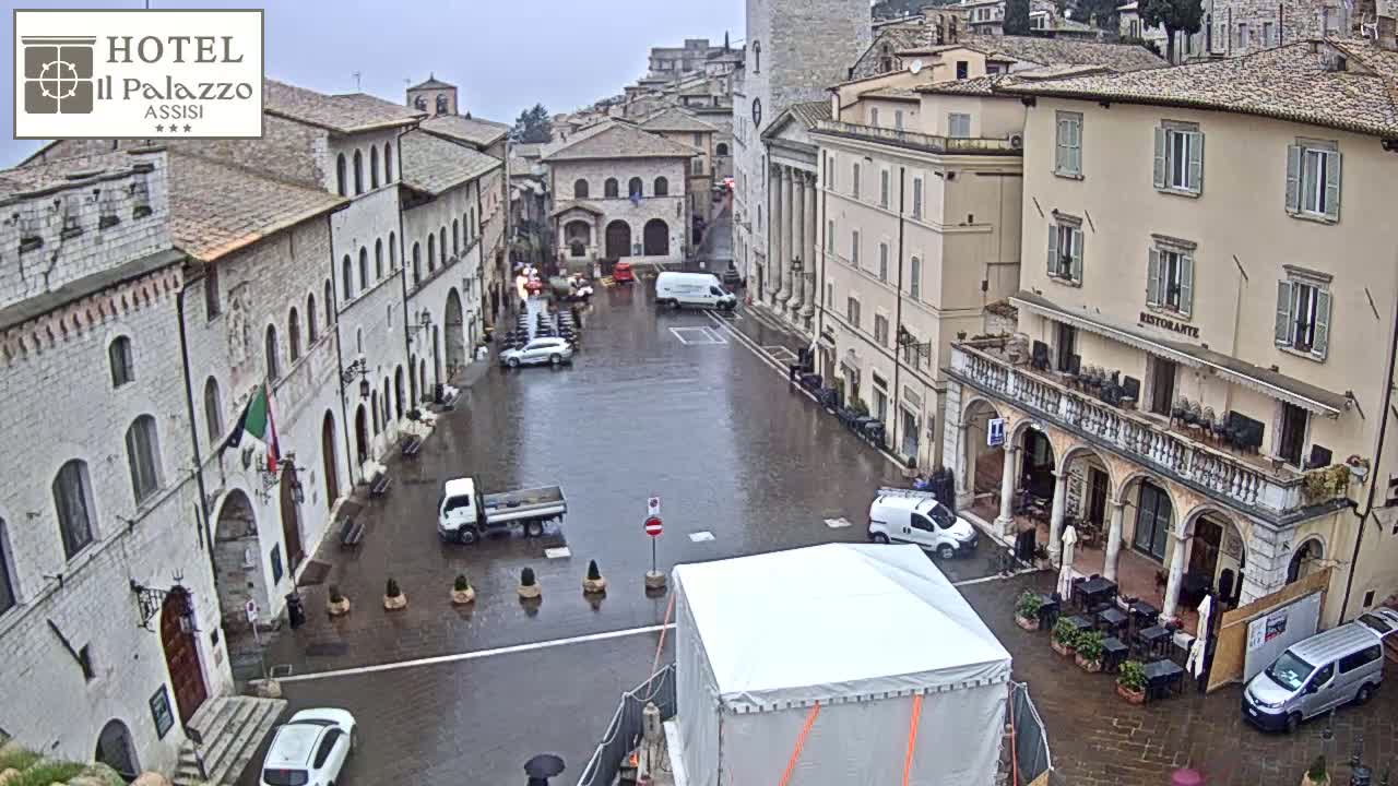 A rain-soaked town square in a European town, surrounded by historical buildings and a few parked vehicles.