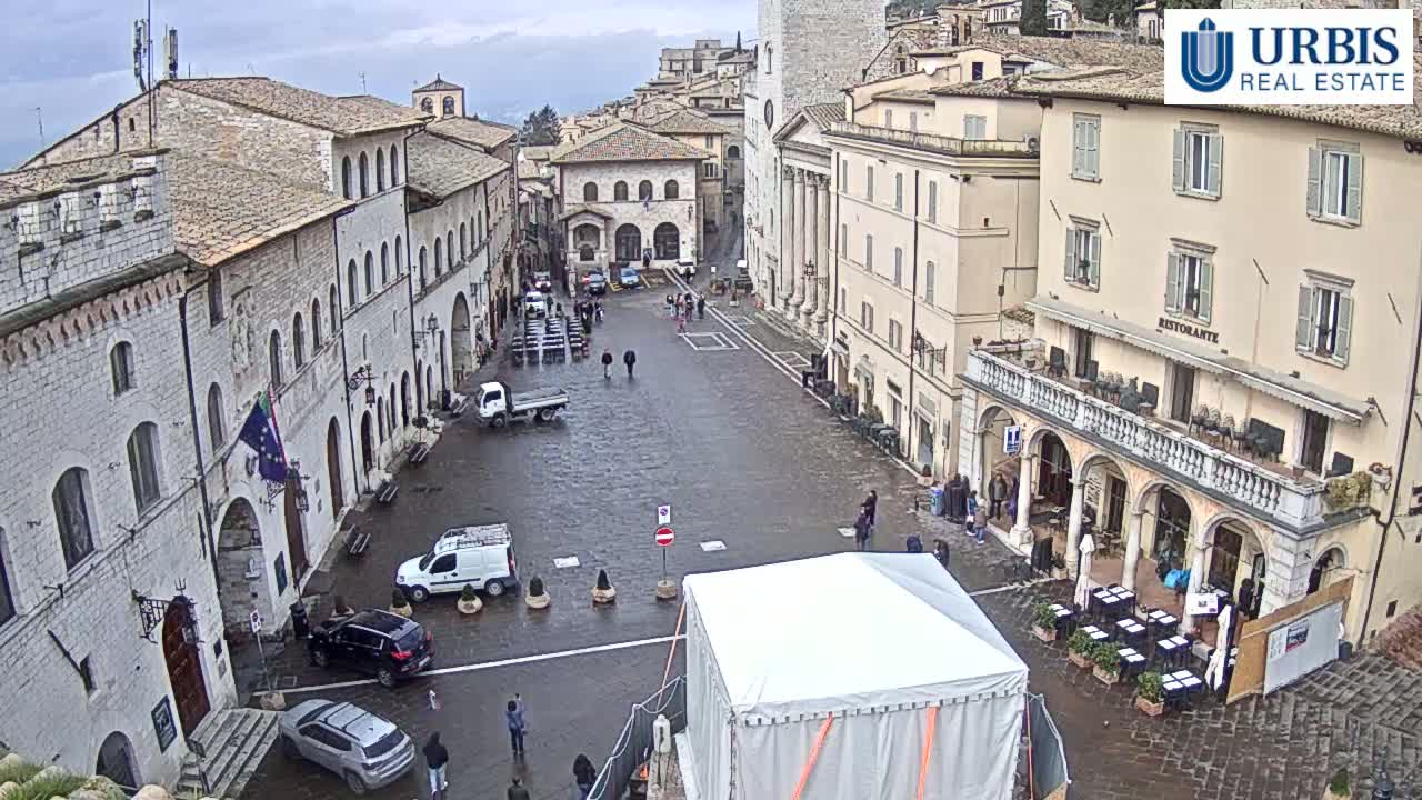 A wet, overcast day in a large, stone-paved town square surrounded by historic buildings, with a few people and vehicles present.