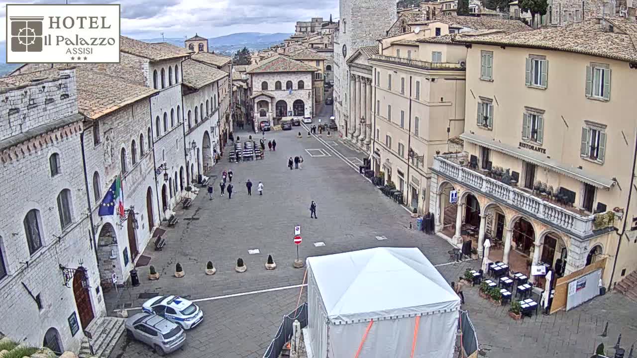A large, mostly empty town square in overcast weather is surrounded by aged stone buildings, with a few people walking around and some cars parked near the bottom, and a white tent in the center.