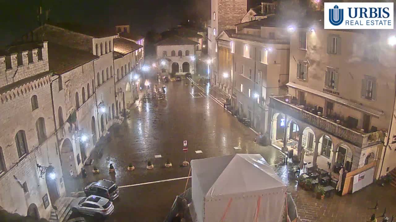A rain-slicked town square at night, surrounded by stone buildings and a few parked cars, with a large tent structure in the foreground.