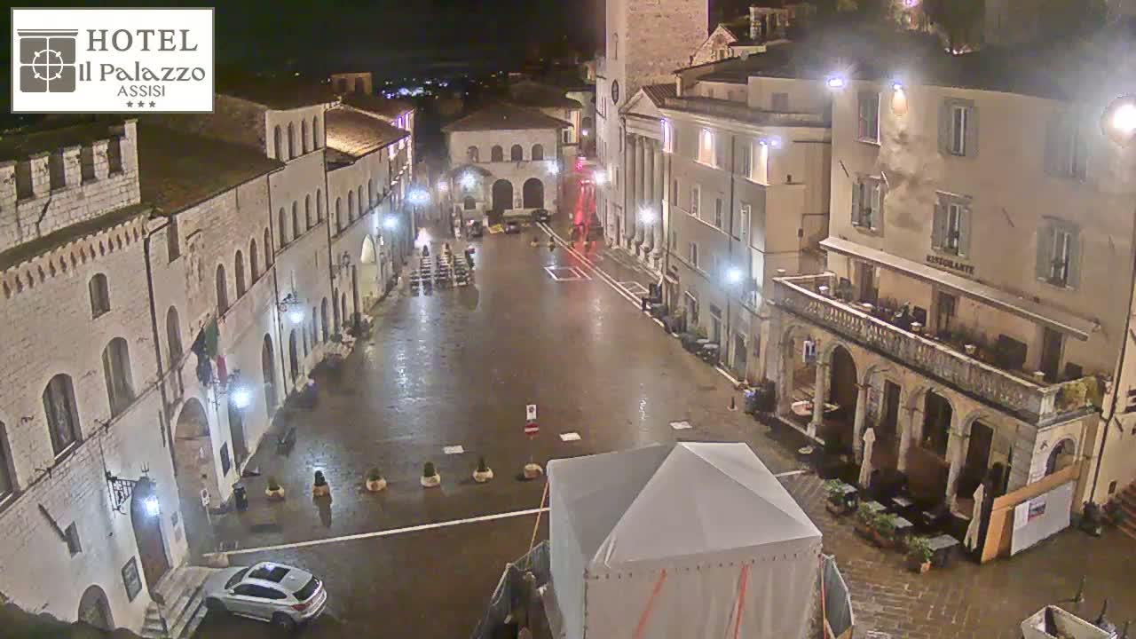 A rain-slicked town square at night, surrounded by historic buildings, with a lone car parked and a large tent in the center.