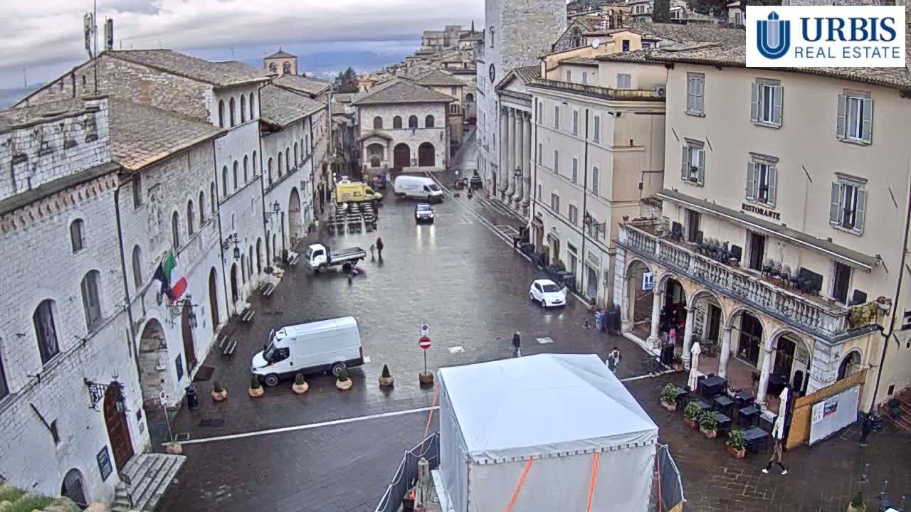 A wet town square in overcast weather is surrounded by several light-colored buildings, with a few vehicles and pedestrians visible.