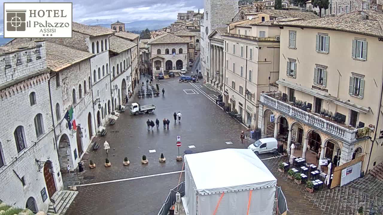A wet, overcast day in a large, stone-paved town square surrounded by historic buildings, with a few pedestrians and parked vehicles visible.