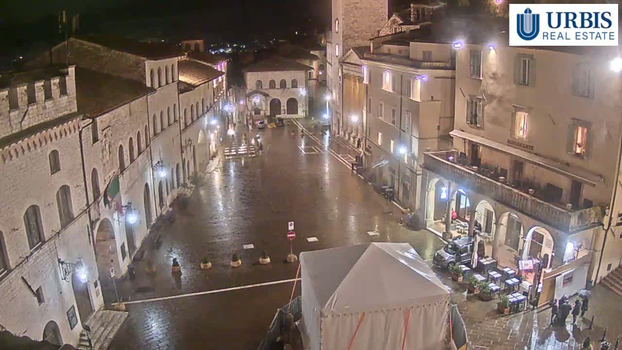 A rain-slicked town square at night, surrounded by historic stone buildings, with a few people and outdoor seating visible.