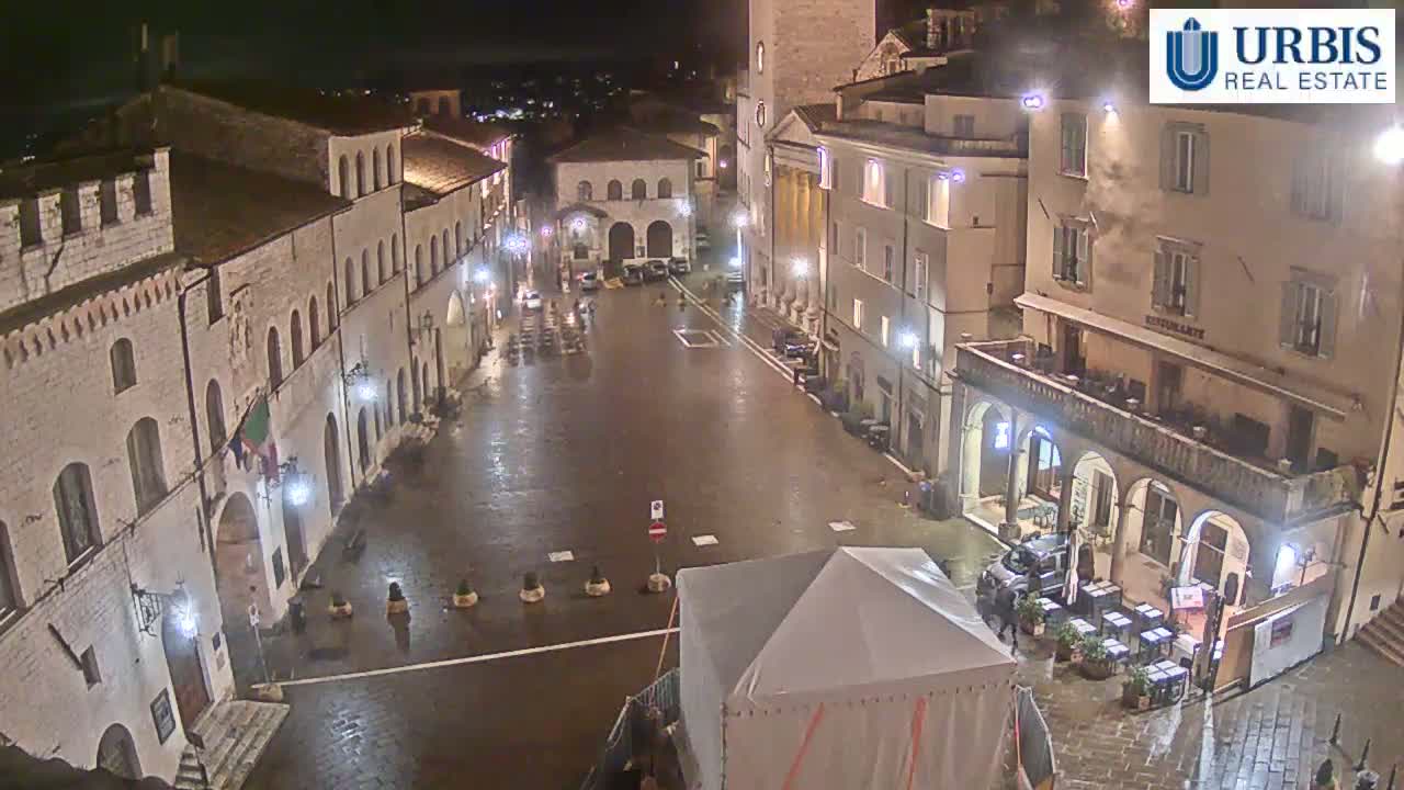 A wet, empty town square at night is surrounded by stone buildings with arched entryways and outdoor seating areas.