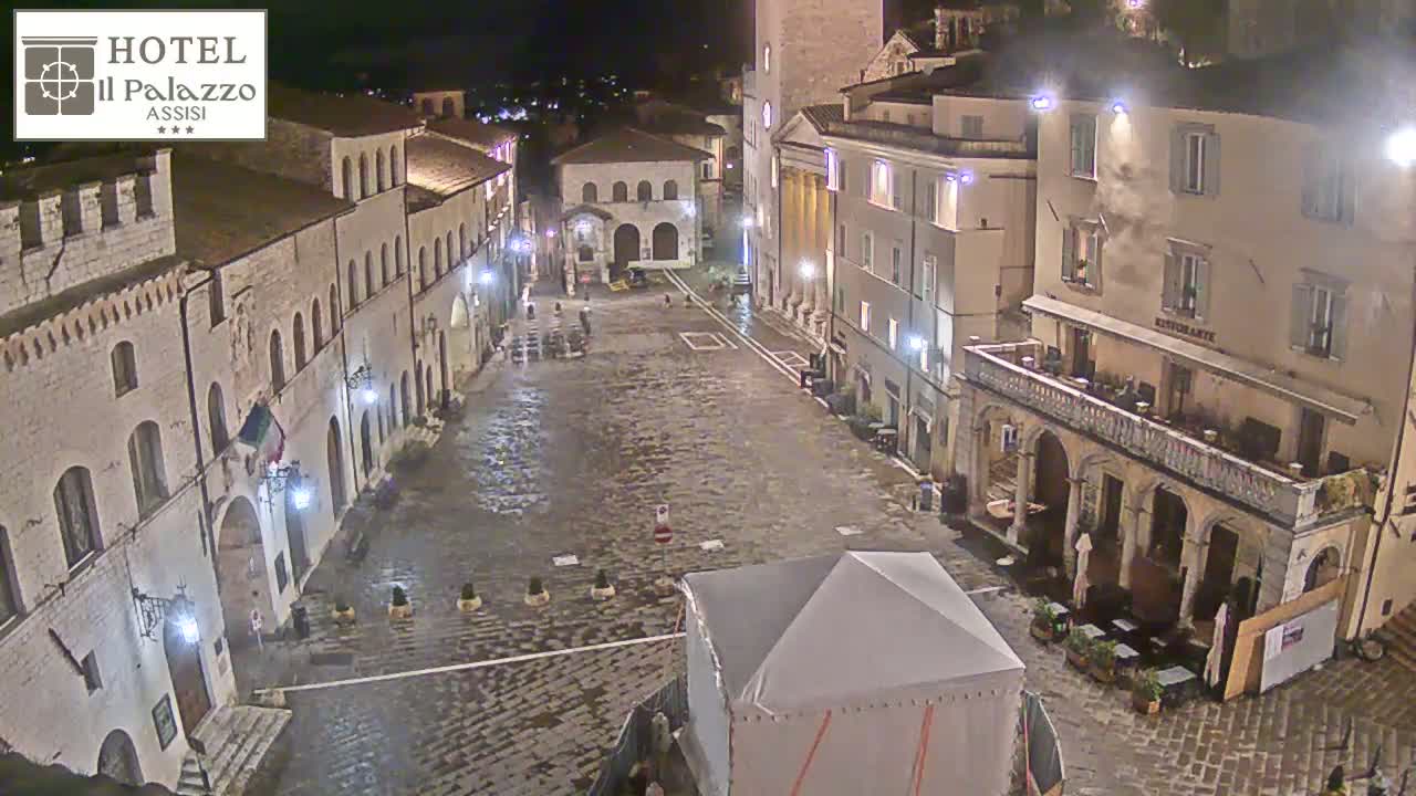 A wet, cobblestone town square at night is surrounded by historic-looking buildings, one of which is partially covered by a temporary tent.