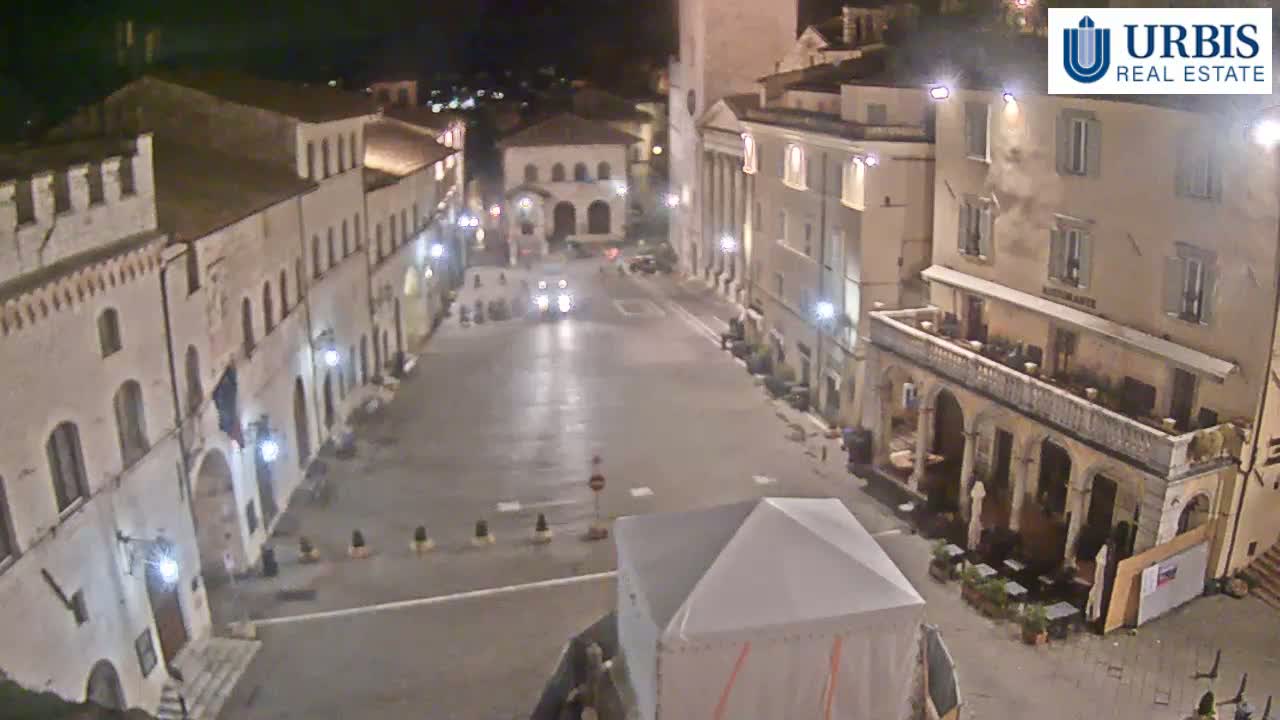 A nighttime, high-angle view of a mostly empty town square surrounded by multi-story buildings, with a few cars visible and a white canopy structure in the foreground.