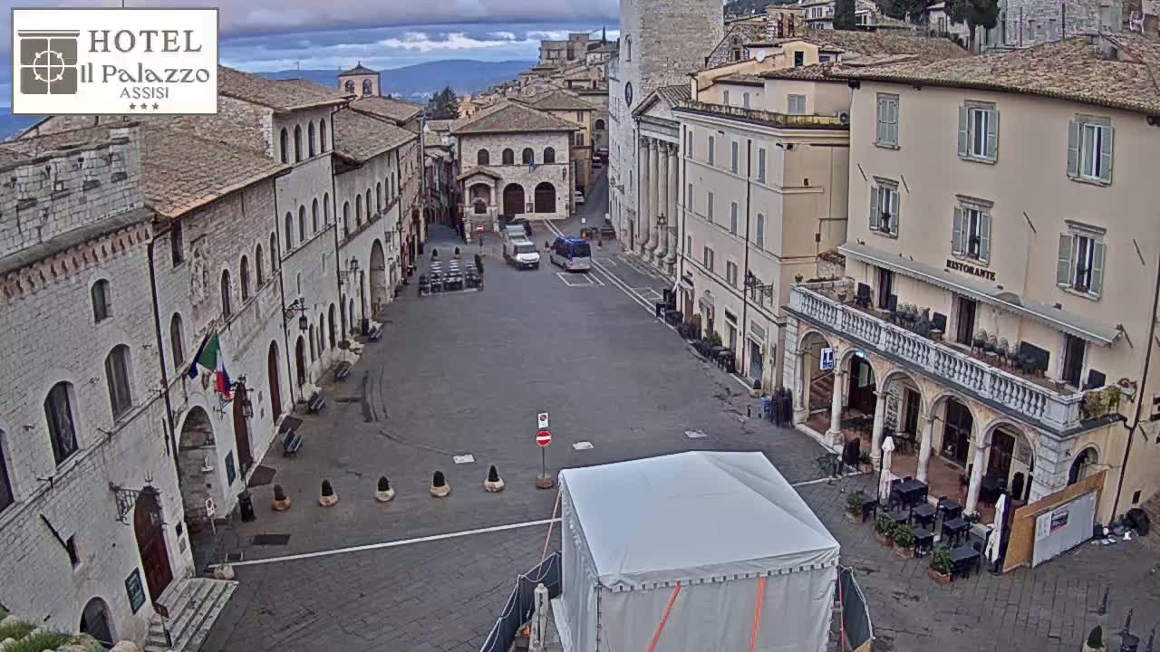 An overcast day reveals a mostly empty town square in a European city, with a few vehicles and a large tent in the center, surrounded by stone buildings.