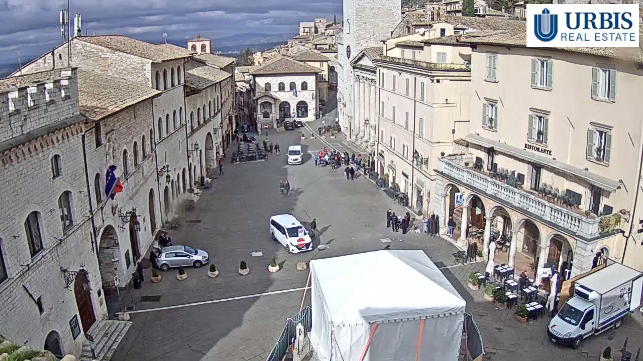 A large town square with several buildings, some cars, and a few people is visible under a cloudy sky.
