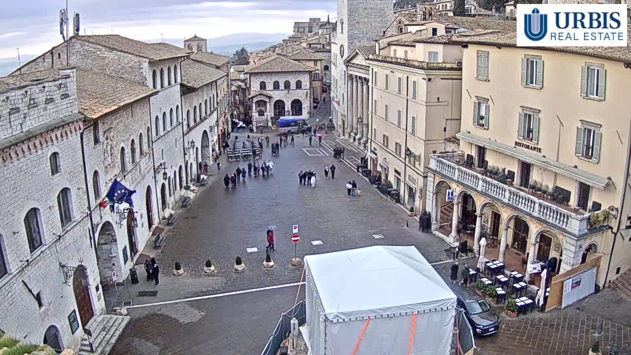 A large, empty town square with several stone buildings surrounding it on an overcast day, with a few people walking around and some outdoor seating set up.