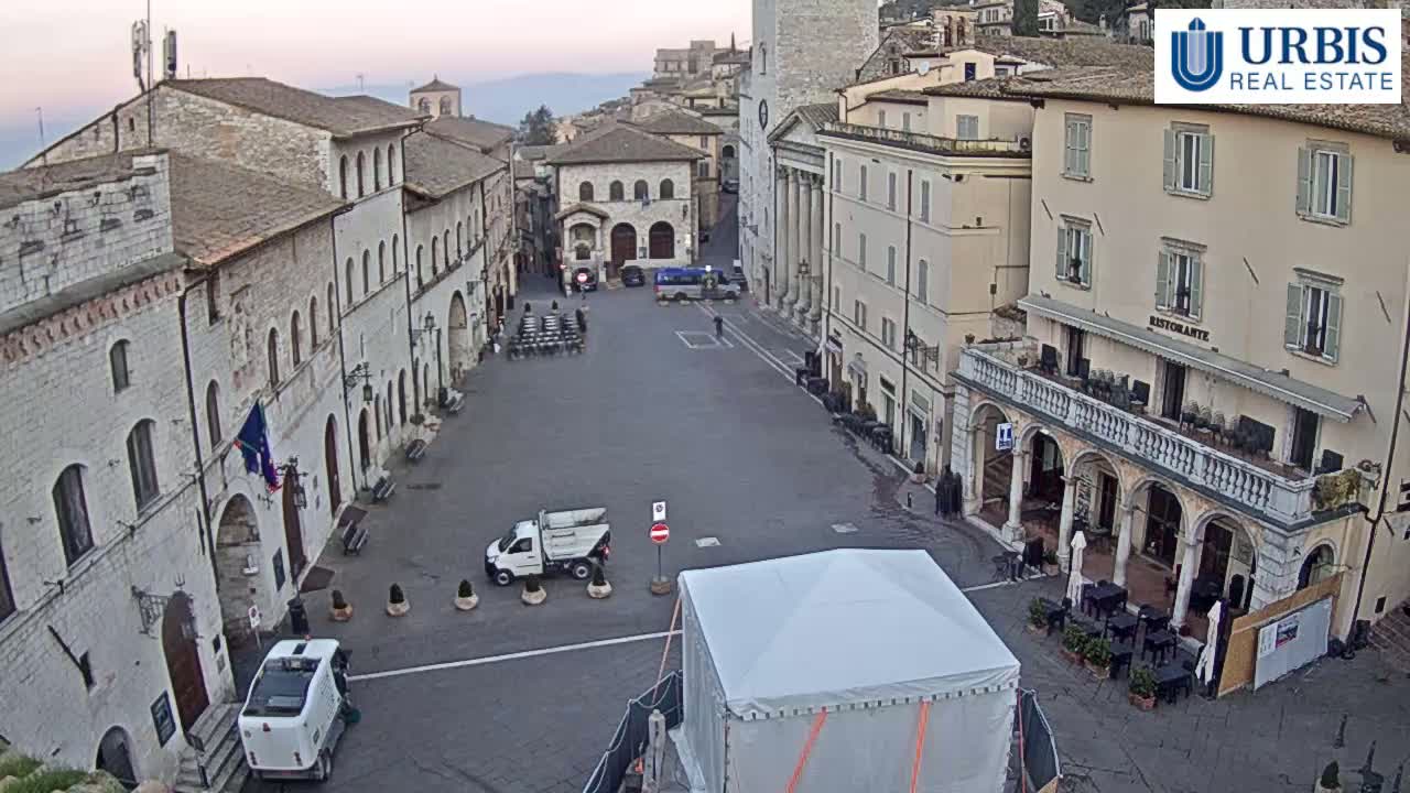 A mostly empty town square in a historic European town, under a pale, possibly sunrise or sunset sky, is surrounded by aged stone buildings, with a few vehicles and a large white tent visible in the center.
