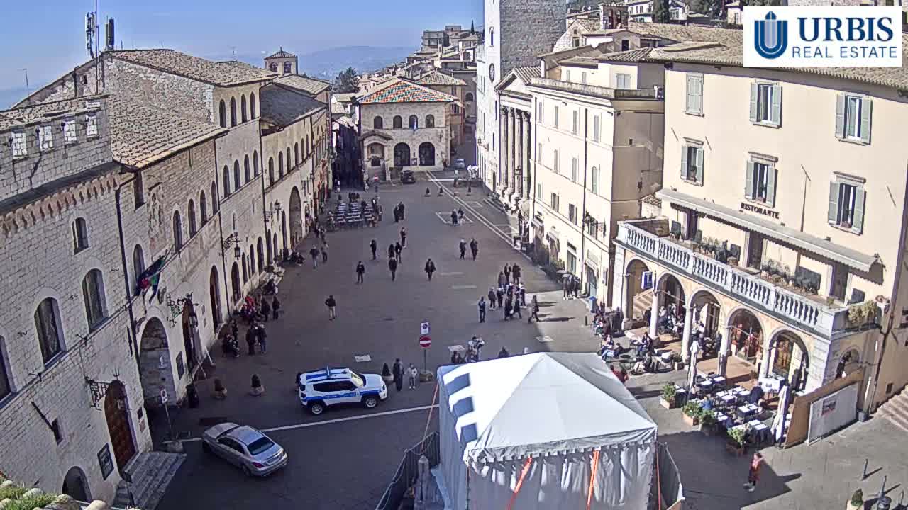 A sunny day in a town square surrounded by historic buildings, with people walking around and cars parked, and a large white tent in the center.