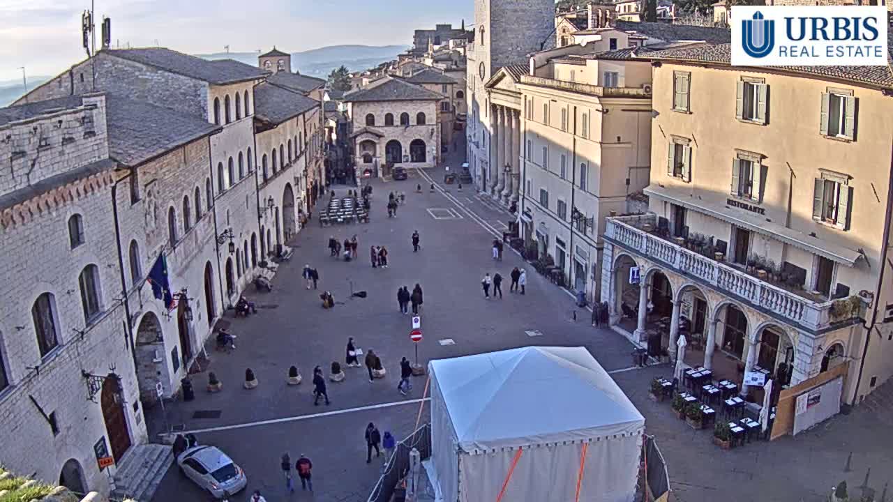 A large, mostly empty town square is surrounded by ancient-looking buildings under a clear, sunny sky.