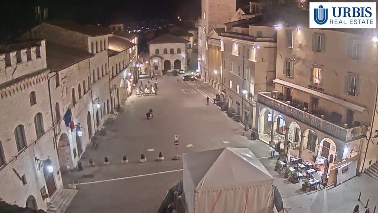 A mostly empty, brightly lit town square at night, surrounded by historic-looking buildings, is shown from an elevated perspective.