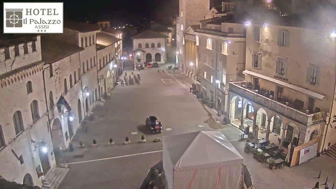 A nighttime, high-angle view of a town square surrounded by historic-looking buildings, with a few cars and people sparsely visible.
