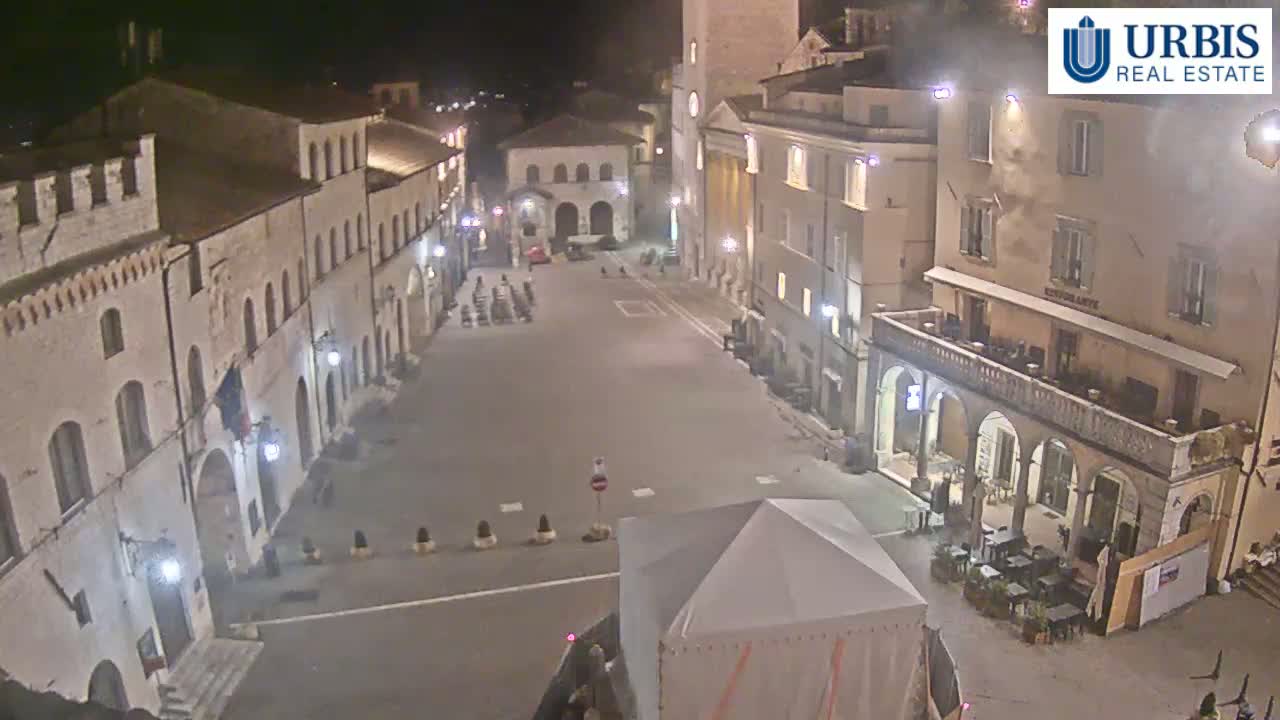 A mostly empty town square at night, surrounded by stone buildings, is illuminated by streetlights.