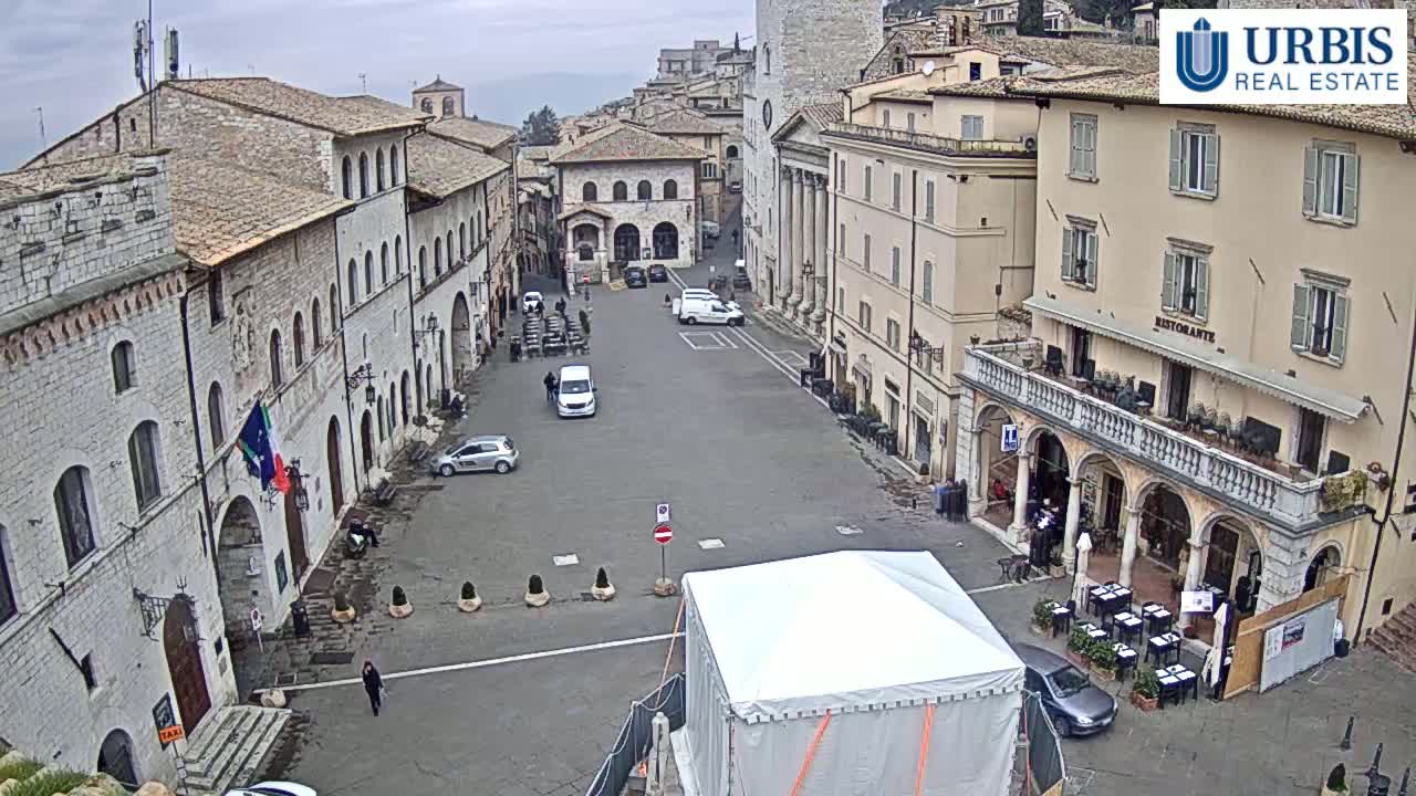 A mostly empty town square in a historic European town, under an overcast sky, features several stone buildings, a few parked cars, and a large white tent in the center.