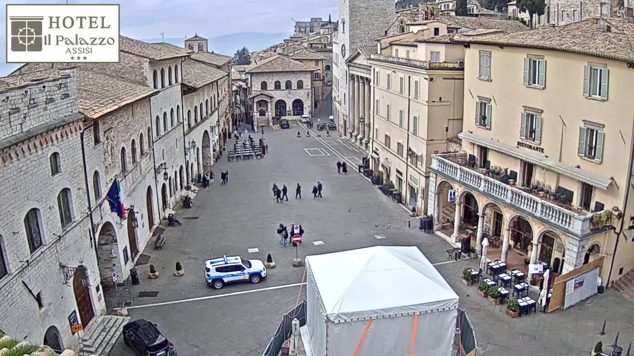 A mostly empty town square in an overcast European town is surrounded by stone buildings, with a few people and a police car visible, and a white tent in the center.