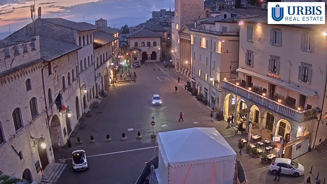 A twilight view of a town square in a European-style town, with several buildings, a few cars, and a small tent in the center.