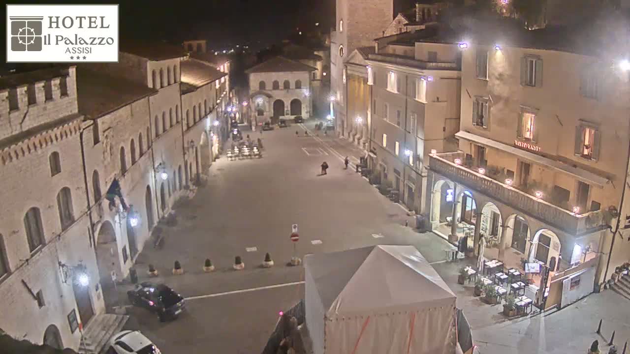 A mostly empty town square at night, surrounded by illuminated buildings, with a few people and cars visible.