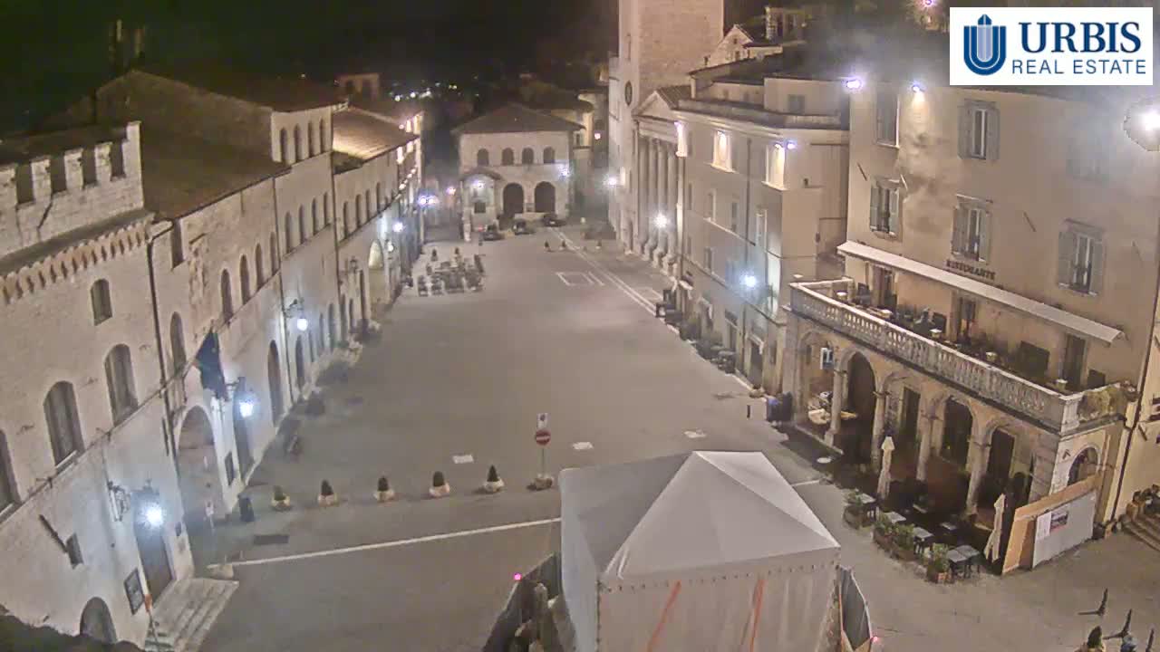 An empty, brightly lit town square at night, surrounded by old stone buildings.