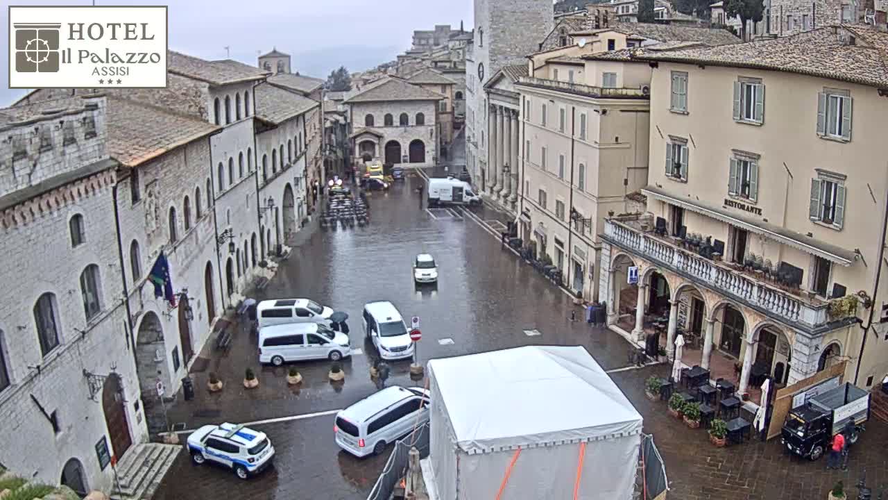 A rain-slicked town square in a European-style town, surrounded by stone buildings, with several vehicles parked and a few people walking about.