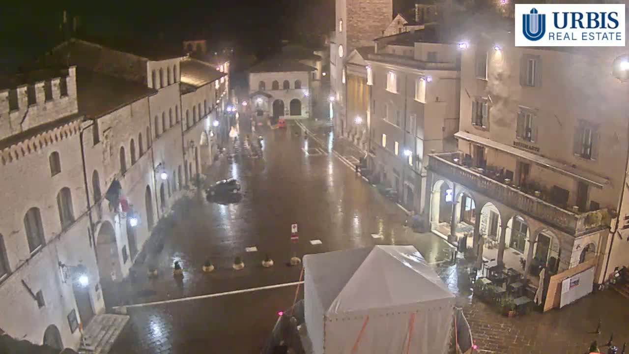 A rain-slicked town square at night, surrounded by historic-looking buildings, with a temporary tent structure in the foreground.