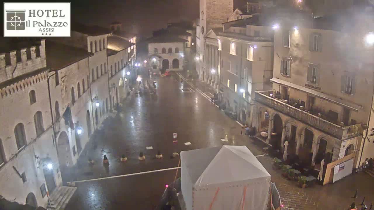 A rain-slicked town square at night is surrounded by historic-looking buildings, with a few people visible and a temporary structure in the center.