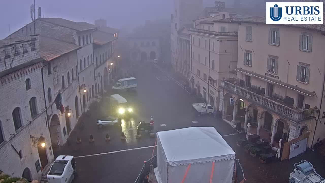 A foggy town square, surrounded by stone buildings, with several vehicles parked and a large covered structure in the foreground.