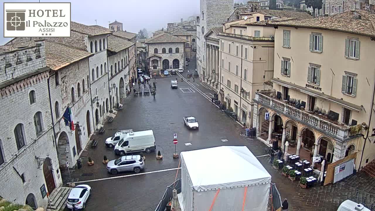 A wet, paved town square is surrounded by stone buildings on an overcast day, with several vehicles and a few people visible.