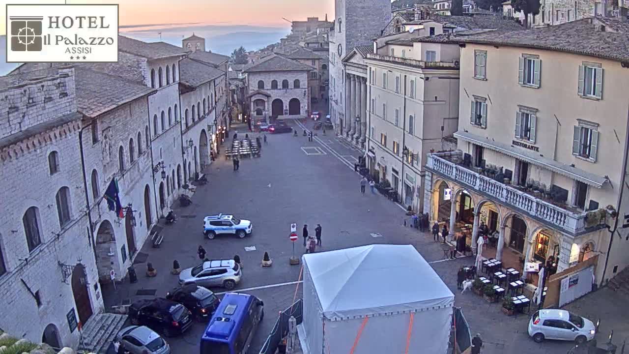 A large, empty town square surrounded by historic buildings at sunset, with several cars and a few people present.