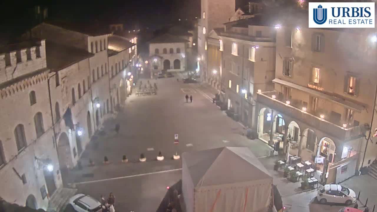 A nighttime, high-angle view shows a mostly empty town square surrounded by illuminated buildings.