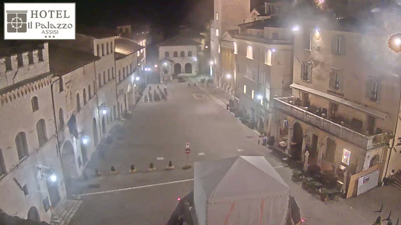 A mostly empty town square at night, surrounded by old buildings, with a few people and a small tent visible.