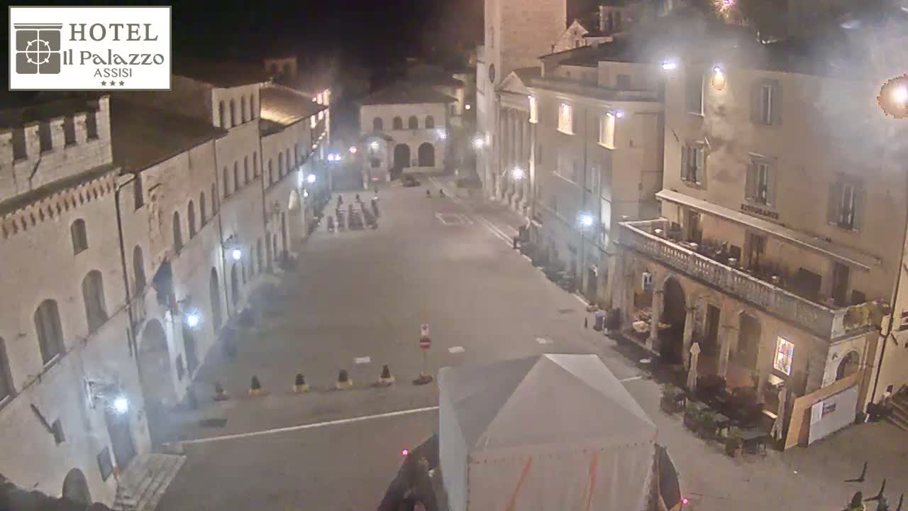 A nighttime, high-angle view of a mostly empty town square surrounded by stone buildings under artificial light.