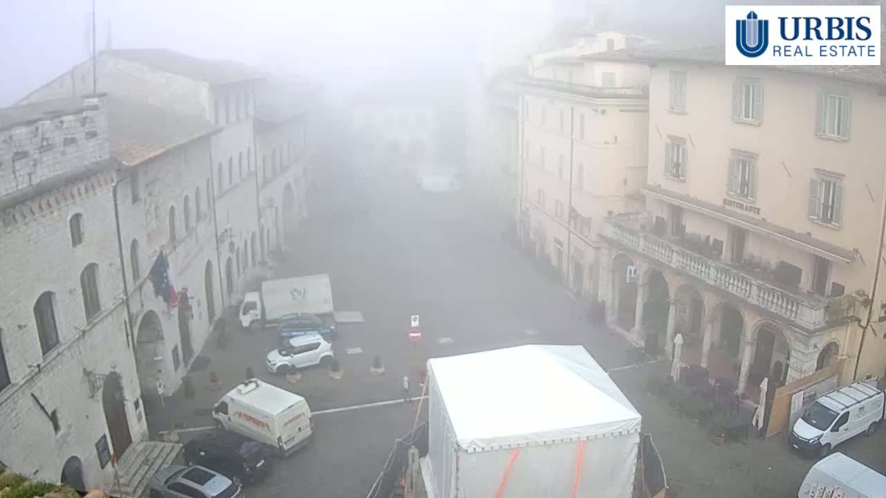 A foggy town square is seen from above, with several vehicles parked and a large white tent in the center.