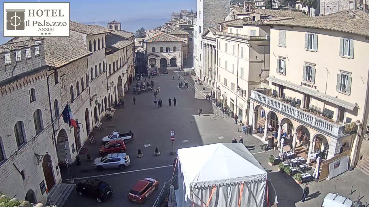 A sunny day reveals a large town square in a historic Italian city, with several parked cars and a few pedestrians, and a large white tent in the center.