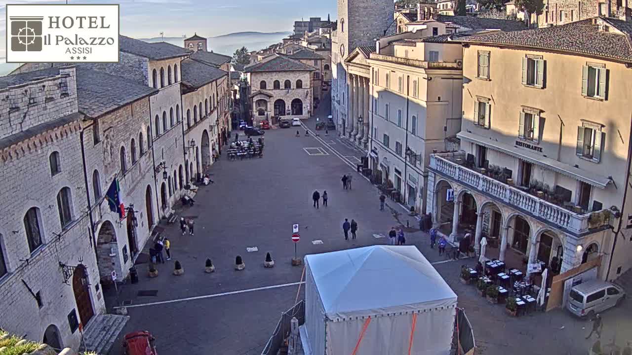 A large, mostly empty town square is surrounded by aged stone buildings under a clear, bright sky.