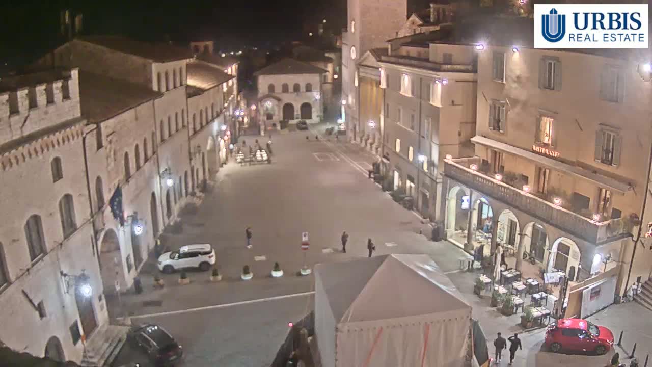 An empty, stone-walled plaza at night is lit by streetlights, with a few people and parked cars visible, surrounded by buildings.
