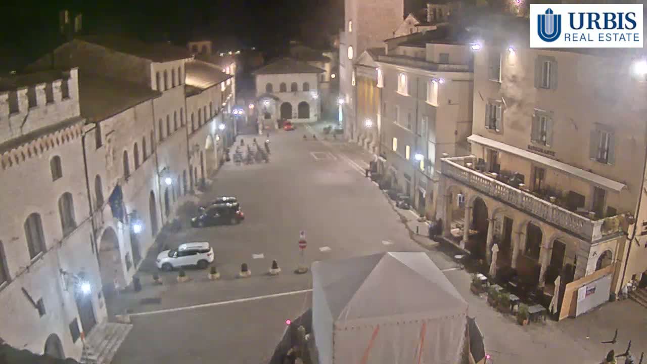 An empty, brightly lit town square at night is surrounded by multiple-story buildings, with a few cars parked nearby.