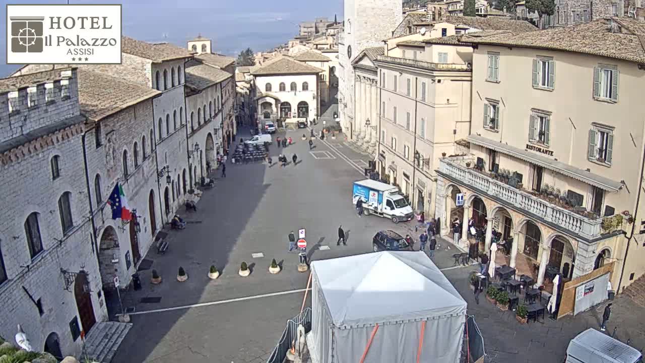 A large, mostly empty town square in bright sunlight is surrounded by historic stone buildings, with a few people and vehicles visible.