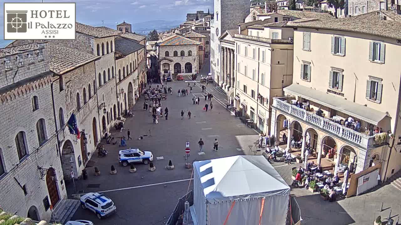 A mostly sunny town square in Italy is filled with people, two police cars, and a white tent; buildings surround the square.
